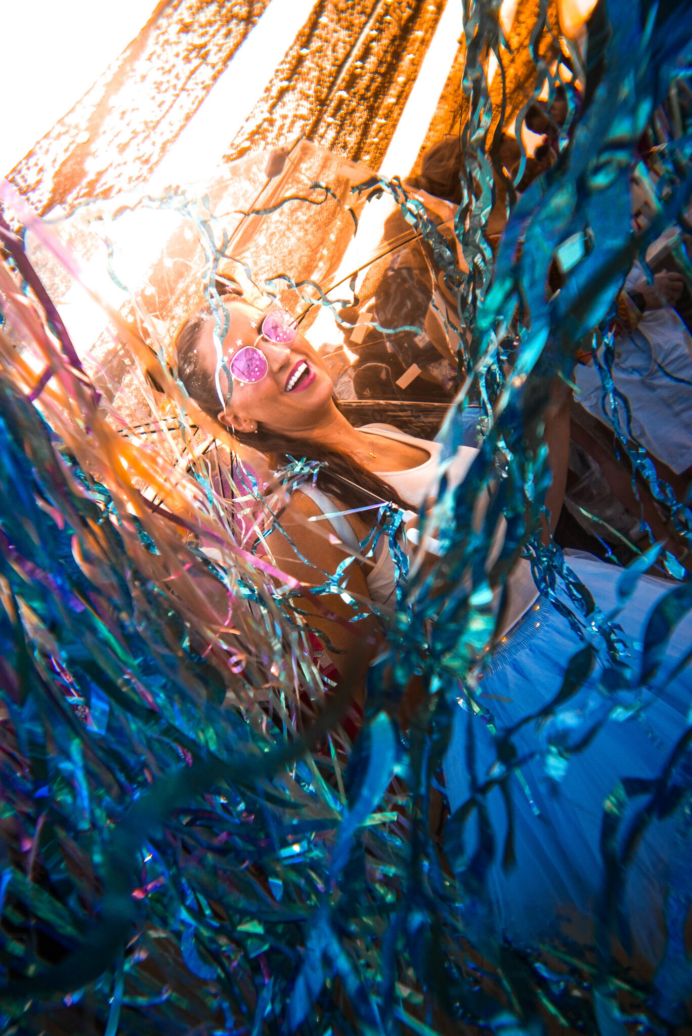 Woman in pink sunglasses smiling and dancing through metallic streamers at an outdoor festival, lit by warm sunlight.