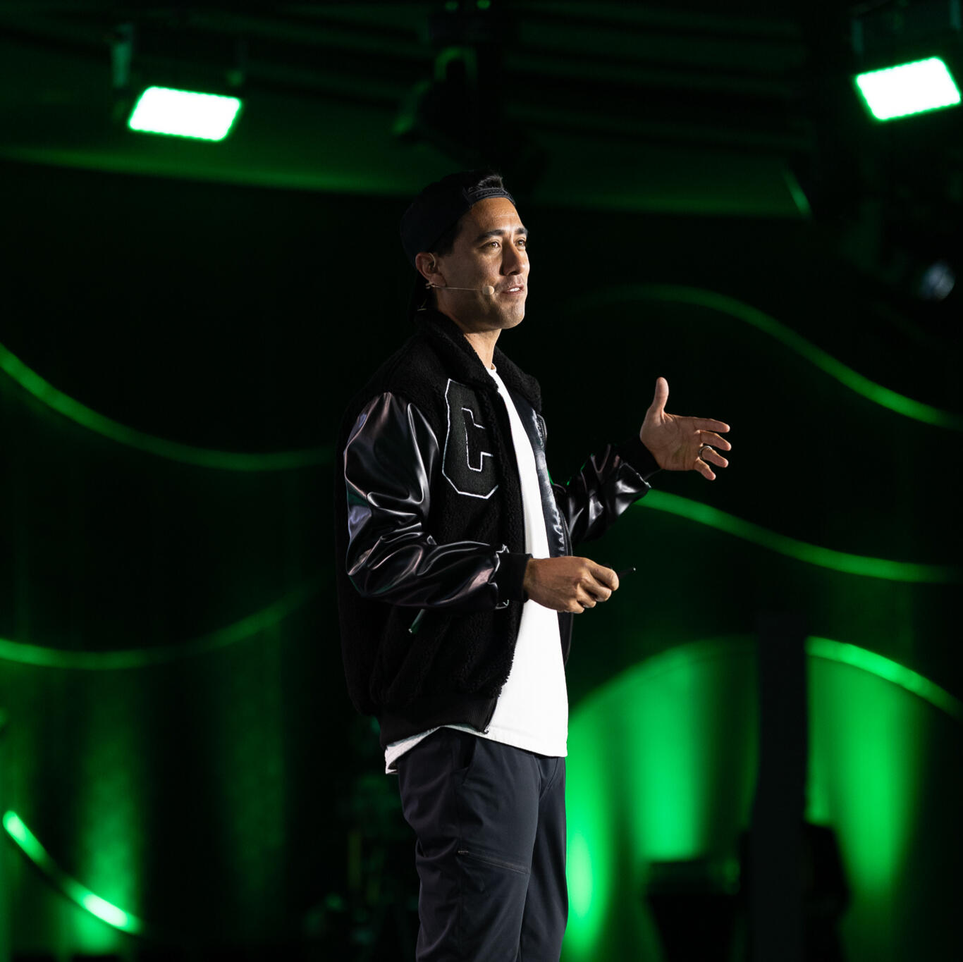 Man wearing a black varsity jacket, white t-shirt, and red sneakers speaks on stage under green lighting at a professional event. He gestures with one hand and wears a headset microphone.